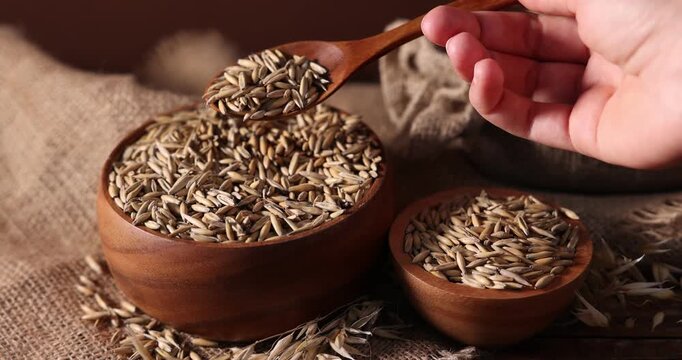Woman adding oat grains from spoon into bowl at table with burlap cloth against brown background, closeup
