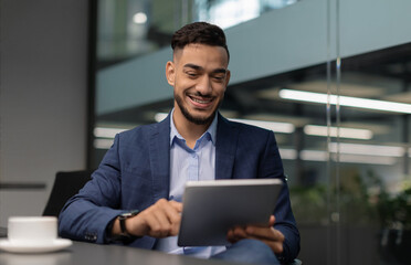 Cheerful arabic millennial guy in nice suit CEO sitting at table, using digital tablet at office,...