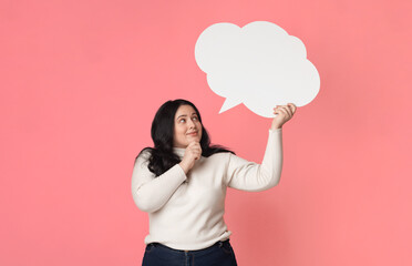Pensive plus size woman holding and looking at blank speech bubble over her head, standing on pink background