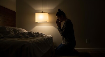 A woman kneeling in prayer beside her bed in a dimly lit bedroom, a low-key, intimate portrait conveying serenity and peace.