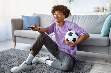 Full length of black teen guy watching football on TV, using remote control, holding soccer ball at...