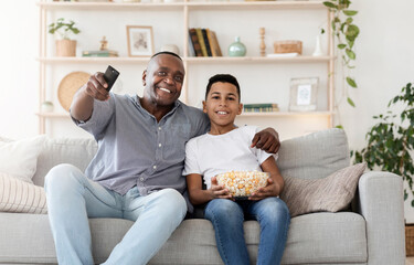 Happy black grandpa and his preteen grandson sitting on couch eating popcorn and watching TV...