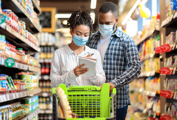Grocery Shopping. Black Couple Wearing Face Masks In Supermarket, Buying Groceries With Checklist, Walking With Trolley In Food Store Indoors. Customers Buy Essentials During Virus Pandemic Outbreak