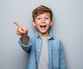 A joyful young boy, wearing a denim shirt, points enthusiastically, showcasing excitement and a bright personality against a neutral backdrop.