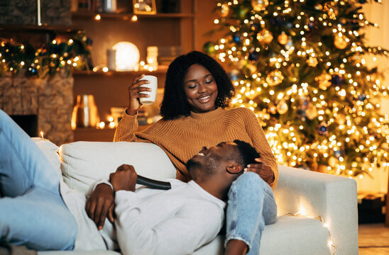 Lovely black woman spending time with her happy boyfriend on Christmas eve in decorated living room, copy space. Happy African American couple cuddling on sofa, drinking coffee on Xmas at home