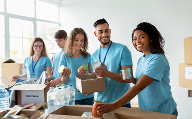Diverse group of volunteers sorting donated food in boxes, working in community charity donation center and smiling at camera, preparing boxes for poor and homeless people