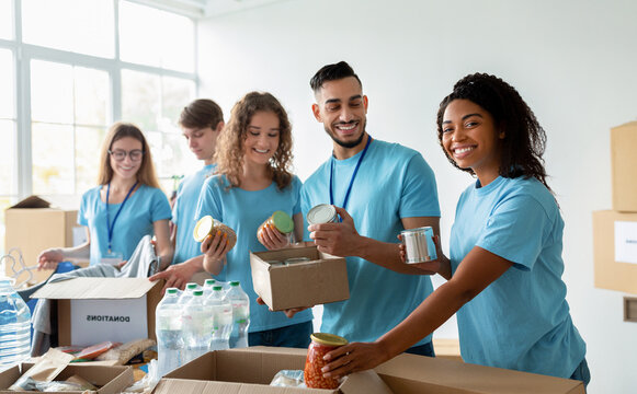 Diverse group of volunteers sorting donated food in boxes, working in community charity donation center and smiling at camera, preparing boxes for poor and homeless people
