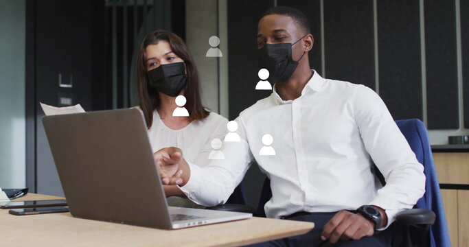 Wearing masks, coworkers in suits pointing at laptop screen on table in conference room