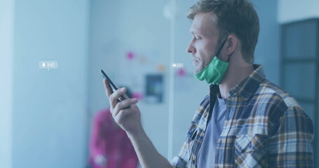 Man with green mask at chin using smartphone in office glass partition, sticky notes, overlay icons