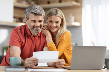 Happy middle aged spouses reading letter from bank, sitting at table in front of laptop, drinking coffee, checking correspondence together, kitchen interior, copy space. Mortgage, loan concept