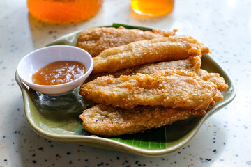 Top view of traditional Asian snack called fried banana served with sweet dipping sauce on a green banana leaf plate on a white table. Suitable for Asian cuisine, tropical desserts, traditional snacks