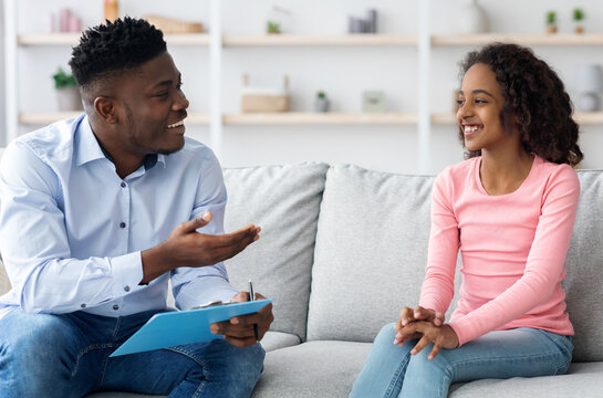 Pretty black girl teenager attending therapy session with psychologist young handsome man, cheerful child psychotherapist having conversation with smiling kid, sitting on couch at clinic