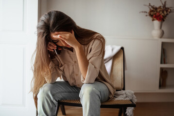 Obraz premium Despaired sad millennial mixed race female calls by phone, talks, sits on chair, suffering from bad news in room interior, free space. Communication remotely, problems, stress at home