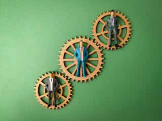 The image depicts a small figurine of a businessman standing on three interconnected wooden gears, set against a plain green background