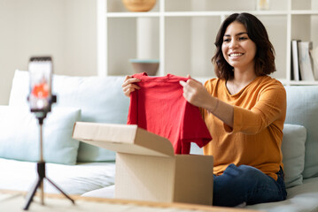 Smiling Arab Female Influencer Unpacking Clothes While Recording Video Content At Home, Middle Eastern Blogger Woman Opening Box With New T-Shirt In Front Of Camera, Using Smartphone On Tripod
