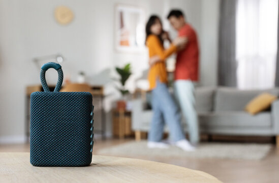 Selective focus on brand new portable speaker over dancing romantic asian couple, blurred background, copy space, home interior. Modern technologies and domestic entertainment concept