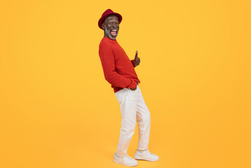 Fototapeta premium Cheerful senior Black man wearing a red sweater and matching hat, giving a thumbs up and looking to the side with a wide smile on a plain yellow background, full length
