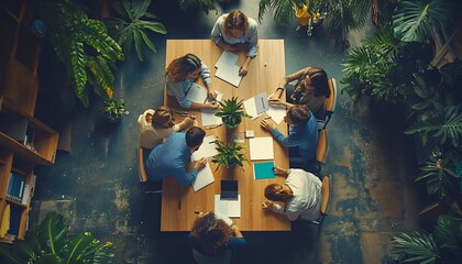 A group of young people are sitting around a large wooden table, working together on projects and surrounded by lush green plants.
