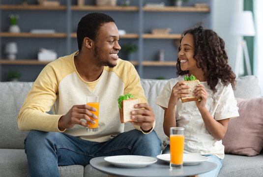 Cheerful african ameican dad and female kid having lunch at home, eating sandwiches and drinking fresh orange juice full of vitamins. Healthy lifestyle, father and daughter, fatherhood concept