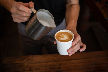 Barista holding a paper cup of latte coffee after pouring steamed milk to making latte art.