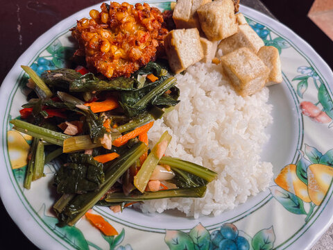 Closed view of Indonesian Food with side dishes of mix vegetables, tofu, frikadel or corn fritters, fried chicken, and steamed rice as main food on white plate.