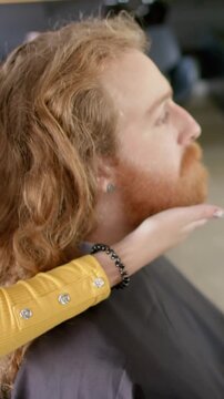 Vertical video: Asian female stylist aligning client's jawline by cupping chin at salon before trim