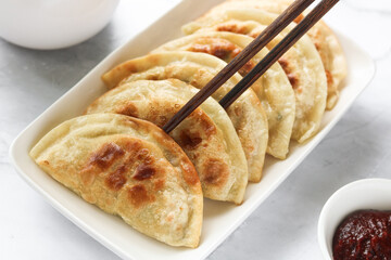 Using chopsticks to eat pan-fried Korean dumplings (Mandu). Beside the plating is a small bowl of spicy red Gochujang sauce.
