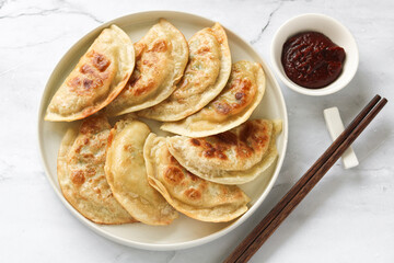 Top view of a plate of delicious pan-fried Korean dumplings (Mandu) arranged neatly with a small bowl of spicy red Gochujang sauce and a pair of chopsticks.