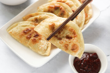 Eating pan-fried Korean dumplings (Mandu) using chopsticks. Beside the plating is a small bowl of spicy red Gochujang sauce.