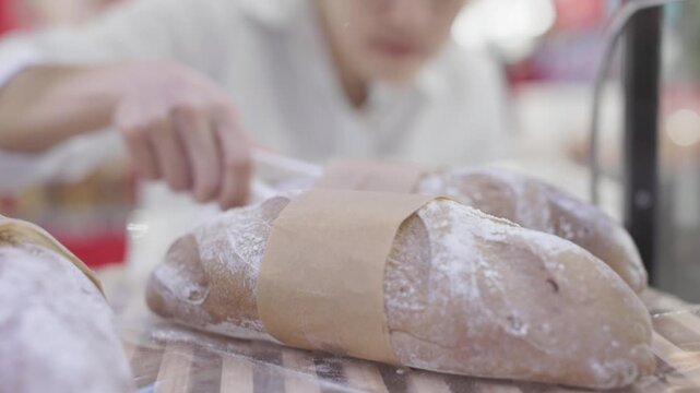 Asian woman using tongs selecting fresh baguette bread in modern supermarket bakery aisle, grocery store food shopping, artisan bread display, healthy lifestyle choice and culinary tradition