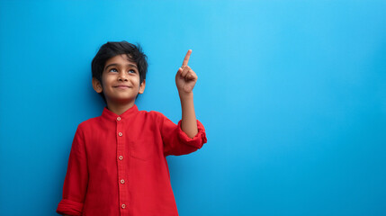 A young boy in a red shirt pointing upwards against a vibrant blue background looking up and smiling