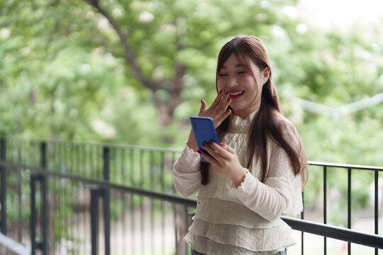 Asian adult woman enjoys smartphone outdoors remote work break happy moment digital communication vibrant green environment urban balcony lifestyle