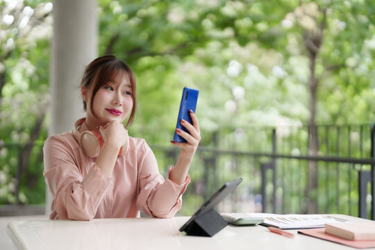 Asian adult woman freelancer works remotely using smartphone tablet outdoors connects digital nomad professional in outdoor cafe green nature park setting