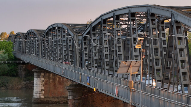 Metallic viaduct spanning Po River, connecting roadway and railway, illuminated by sunset lamps against dramatic twilight sky in Cremona, Italy
