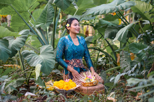 Woman in blue kebaya sitting in jungle with offerings