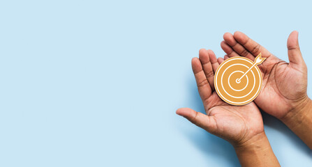Hands holding golden target with arrow symbol on light blue background, representing business...
