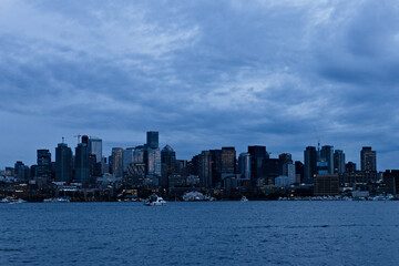 Naklejka premium City skyline at dusk with skyscrapers reflecting evening light under dramatic clouds, viewed across the water, capturing an urban waterfront scene in a moody atmosphere.