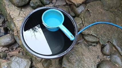A black bucket filled with water with a blue ladle floating in it. Beside the bucket are wet rocks...