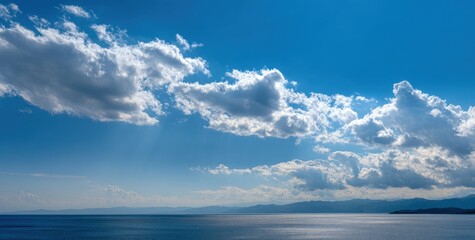 Obraz premium Panoramic view of a serene lake under a vibrant blue sky dotted with fluffy clouds. Horizon line displays distant, indistinct mountains
