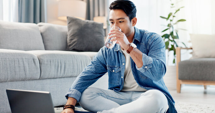 Laptop, drinking water and man in home for remote work, reading report or news update on floor. Computer, thirsty freelancer and glass beverage in lounge, review headline and article for journalism