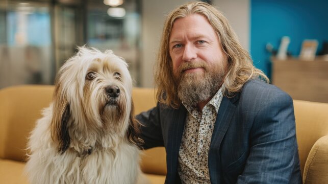 A man with long hair and a well-groomed beard is seated on a couch in a contemporary office. Beside him, a fluffy dog sits calmly, adding a touch of warmth to the environment.