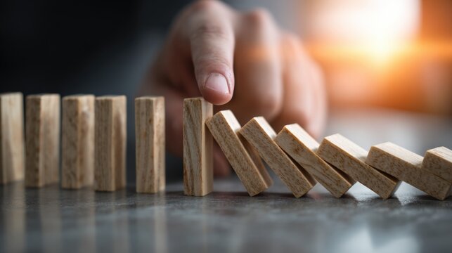 A hand is positioned near a row of wooden dominoes, halting their fall in a workspace. The warm afternoon light creates a serene atmosphere as the dominoes stand in a precarious line