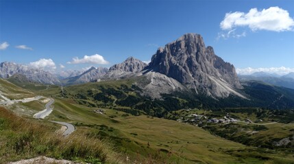 Obraz premium Majestic Dolomites rise against an azure sky with scattered clouds. Winding road leads through lush green hills and quaint village nestled below the peaks.