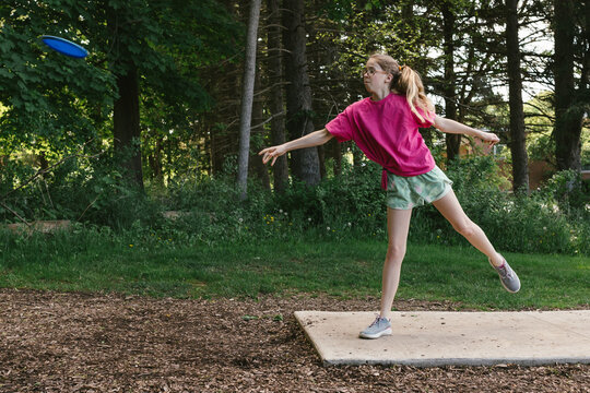 Girl throws disc off tee box in forest while playing disc golf