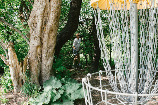 Teenage boy throws disc at golf cage from the forest of green trees