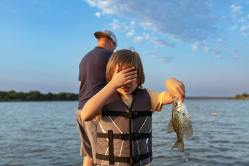 Boy holding fish while covering face on boat at lake at sunrise
