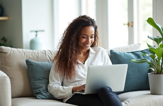 Smiling Black woman with long curly hair wears earphones, works on laptop sitting on sofa. Smart casual attire, relaxed posture suggest comfortable home office environment. Engaged with computer,