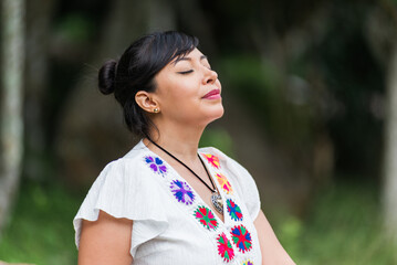 A woman wearing a white embroidered blouse stands outdoors with her eyes closed.