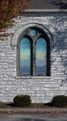 Fototapeta premium Arched window on stone building, reflecting sky in its patterned glass, flanked by shrubbery and autumn leaves