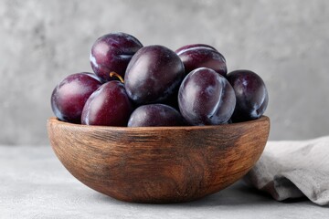 Obraz premium A dark-wood bowl overflowing with plump, dark-purple plums rests on a grey surface, next to a linen napkin